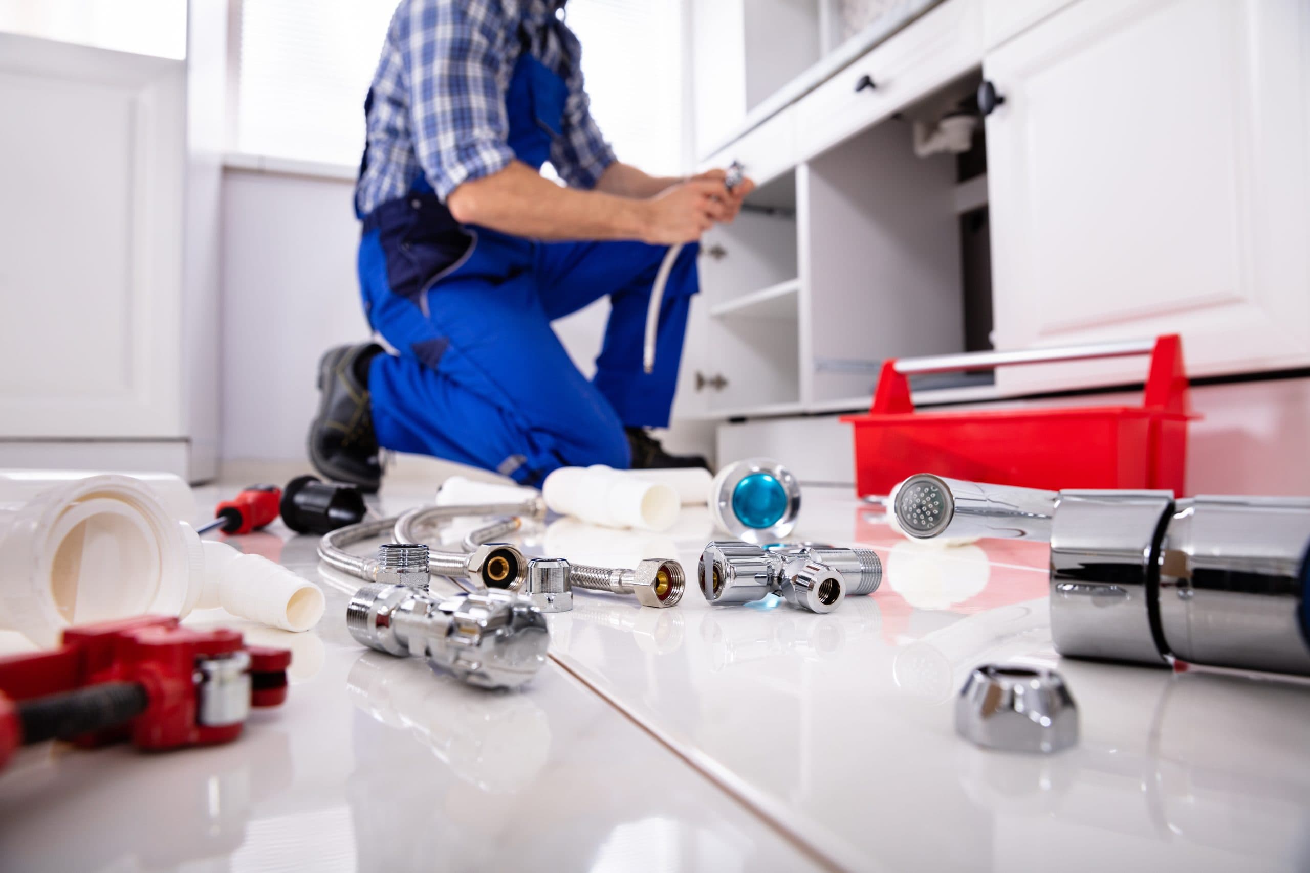 Plumber in blue uniform working under a kitchen cabinet with chrome fittings and tools on tile floor